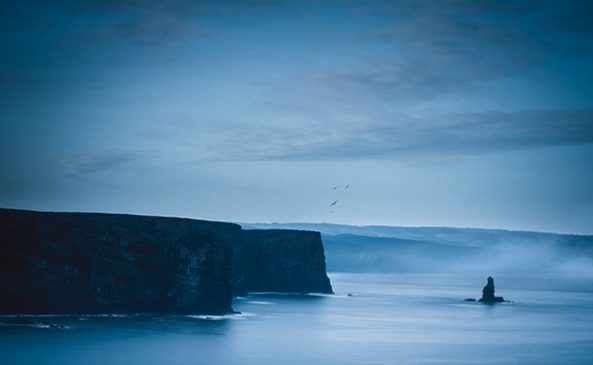 Arrifana at dawn is a dramatic place, with sheer cliffs stretching out into the calm sea and solitary rock formations emerging from the water. The sky and water share almost the same shade of blue, while seagulls fly over the top of the cliffs adding a dynamic element to the tranquil and solitary scenery.