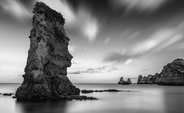 This monochromatic photograph captures a coastal scene. Dominating the foreground is a large, rugged rock formation emerging majestically from the smooth, reflective surface of the water. The sky is filled with soft, wispy clouds, adding a sense of movement and depth to the composition.