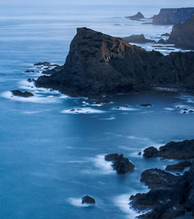 The rugged coastline at Arrifana, Portugal is famous for the dramatic cliffs jutting out into the sea, with various rocky outcrops scattered across the water. The ocean's surface is smooth and has a serene quality, with gentle waves lapping against the rocks. The deep blue tones of the water contrast with the darker hues of the cliffs, creating a tranquil yet imposing atmosphere.