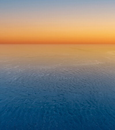 This image captures an ethereal sunset over a vast, calm body of water. The horizon divides the image, where the gradient of the sky shifts from a deep, rich orange to a light blue, eventually blending into a soft, deep blue. The water has gentle ripples, reflecting the subtle hues of the sky.