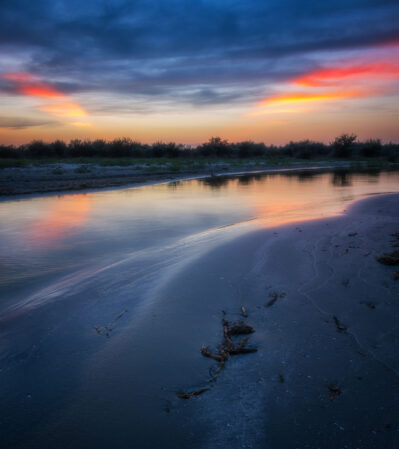 The sun setting below the horizon in the Danube Delta turns the sky into a vivid canvas of star shaped rays of deep blue and rich purple hues, interspersed with striking bands of orange and pink. The riverbank is in shadow, with gentle curves leading the eye towards the horizon, bordered by the dark silhouettes of reeds.