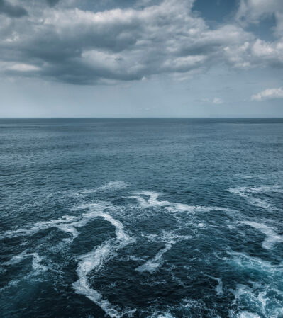 This photograph captures the vastness of the deep, dark blue Atlantic ocean near Sintra, Portugal under a sky filled with thick, gray clouds. The ocean water is dynamic, with white crests indicating gentle waves and swells. The distant horizon line is the meeting point between the sea and the sky, creating a sense of depth and infinity.