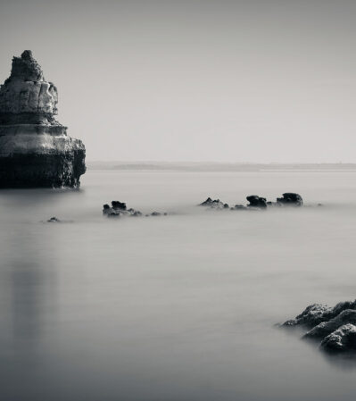 This photograph captures a tranquil coastal scene in monochrome, one of the many secluded beaches near Lagos, Portugal. A large, rugged rock formation stands prominently in the calm, mist-like water, which creates an ethereal atmosphere. The horizon line is faintly visible, giving the image a minimalist and serene quality.