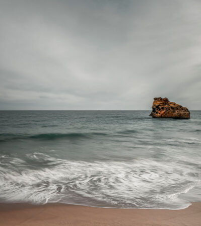 This is a long exposure seascape of a solitary rock projected onto a dark and moody sky. Gentle waves brush against the sandy beach creating a tranquil, moody ambiance to the scene. The rock formation has a rugged texture, providing a focal point against the expansive backdrop of the sea and sky. This image was captured at Praia da Rocha, in the Algarve, after a spring storm.