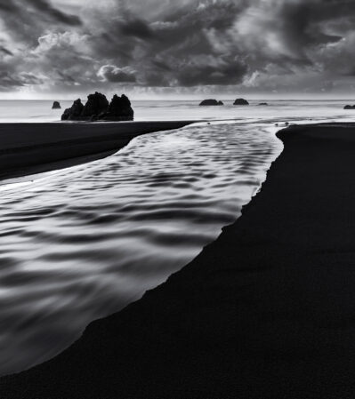 A small stream that formed on Bandon Beach allows the salty waters of the Pacific ocean to flow onshore. In the background, several rock formations rise from the water, their dark silhouettes contrasting against the misty horizon. Above, dramatic cloud formations stretch across the sky, adding texture and depth to the scene.
