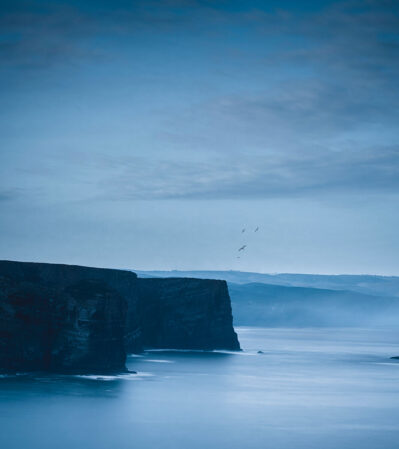 Arrifana at dawn is a dramatic place, with sheer cliffs stretching out into the calm sea and solitary rock formations emerging from the water. The sky and water share almost the same shade of blue, while seagulls fly over the top of the cliffs adding a dynamic element to the tranquil and solitary scenery.