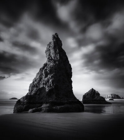 Bandon Beach, Oregon is a dramatic coastal landscape. Central to the composition is a towering rock formation emerging from the shallow waters of the shoreline. The contrast between the dark, jagged rocks and the smooth, reflective surface of the water beneath them creates an evocative and tranquil scene.