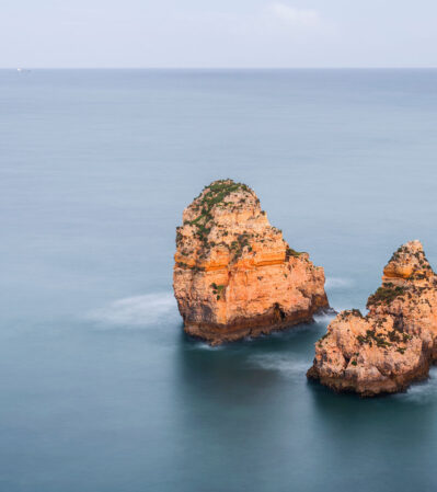 The sun kissed sea stacks emerging from the ethereal, soft, turquoise - blue water near Ponta da Piedade create a beautiful contrast .The horizon is almost blending with the sky, emphasizing even more of the calm and relaxing scene, dominated by a sense of isolation and natural beauty.