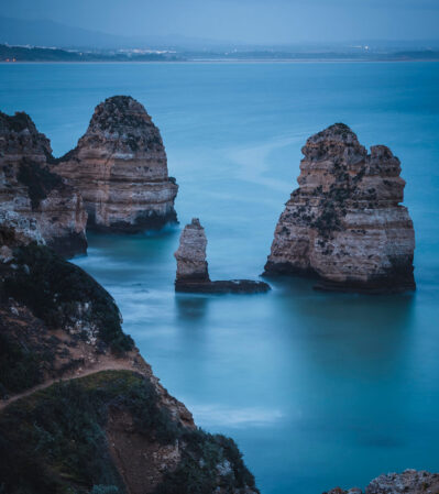 These rugged, weathered rocks at Ponta da Piedade stand majestically against the gentle waves, their sheer surfaces exposing layers of geological history. The distant horizon is subdued, blending seamlessly with the misty mountains and sky, creating an overall harmonious and tranquil composition.