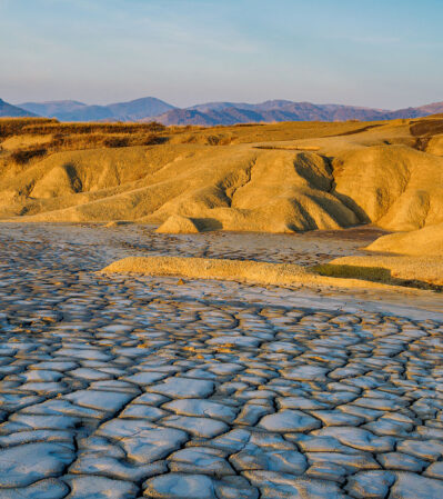 The Mud Volcanoes Natural Park may seem like a parched and desolate environment. The landscape is in a continuous transformation as the mud keeps erupting from small cones, drying up into layers of cracked mud. Golden, grassy rolling hills dominate the midground and a line of distant, muted mountains add depth to the scene in the background.