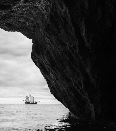 This image combines elements of natural geology with maritime history, creating a powerful composition. Dominating the right side is a massive rock formation that extends vertically, and in the distance, a classic sailing ship sails across a calm, open sea under an overcast sky.