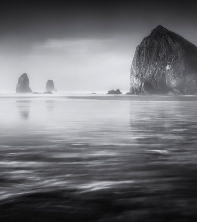 The morning light on Cannon beach, Oregon, creates a serene and ethereal atmosphere. The Haystack rock is imposing, being counterbalanced by the smaller rocks called Needles. The photograph's long exposure creates a smooth and reflective water surface. A subtle crescent moon in the upper right corner provides a delicate accent to the composition.
