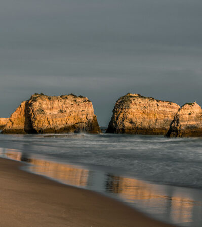 This serene coastal scene of a moody overcast sky in contrast with the illuminated rocks rising from the ocean at Praia da Rocha in Portimao, Algarve, bathed in a warm, golden light, casting reflections onto the wet sand below captures the feeling of peace after the storm.