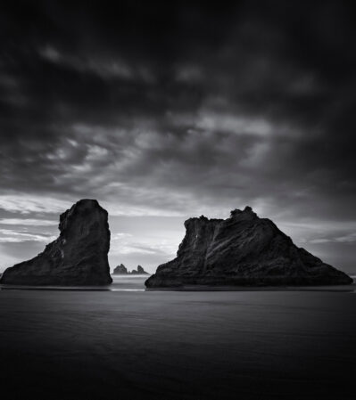 Bandon Beach is peppered with sea stacks of various sizes and shapes. Their rough textures contrast against the smooth, wet sand. In the background, a moody sky with dramatic cloud formations adds depth and a sense of atmosphere to the scene.