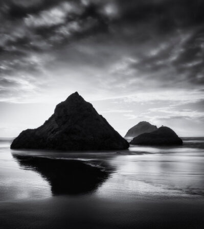After the storm, Bandon beach turns into a serene seascape featuring large rocks emerging from the shallow waters near the shore. The sky is dramatic, filled with clouds that enhance the contrast and mood of the image. The interplay between the rugged rocks, the tranquil water, and the striking sky creates a powerful and evocative image.