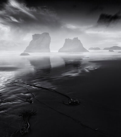 The Oregon coast after the storm becomes a dramatic landscape with large, rugged rock formations emerging from the ocean and fog rising from the water. In the foreground, seaweed and small water channels create textures on the sandy shore, leading the viewer's eye towards the imposing rock structures in the distance.