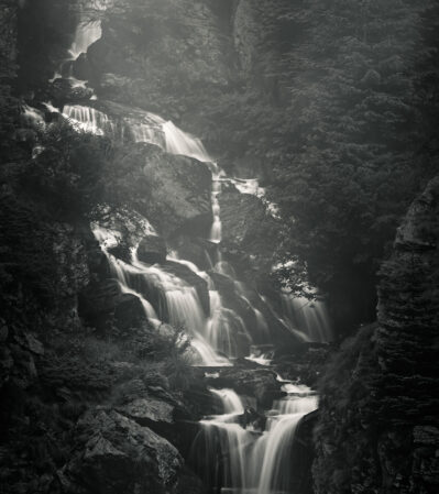 This black-and-white photograph captures the majesty of a cascading waterfall amidst a forested landscape in the Fagaras Mountains of Romania. The mist rising from the waterfall adds an atmospheric depth to the scene, enhancing the sense of tranquility and natural beauty.
