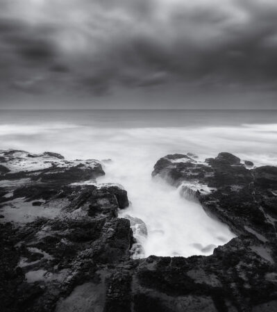 This photograph captures the rugged, rocky coastline of Oregon at Yachats. The long exposure effect creates a silky, ethereal quality in the water, contrasting with the dark, sharp textures of the rocks. The horizon is positioned towards the upper third of the frame, dividing the tumultuous, cloudy sky and the serene, misty ocean. Cape Perpetua is famous for its dramatic and moody natural beauty.