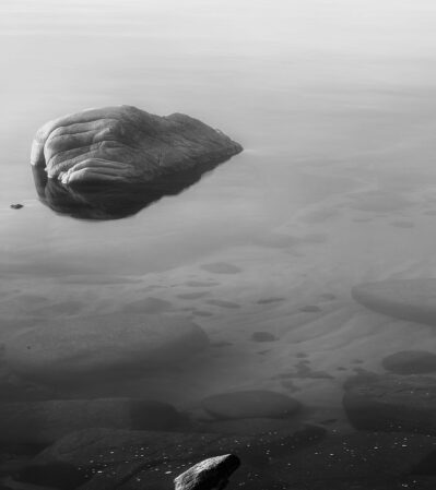 The black and white photograph captures a calm day at Lake Tahoe. The focal point of the image is a large rock partially visible above the smooth surface of the water, creating a reflection. The photograph emphasizes the textures and contrasts of the natural elements, and the monochromatic tones evoke a serene and meditative atmosphere.