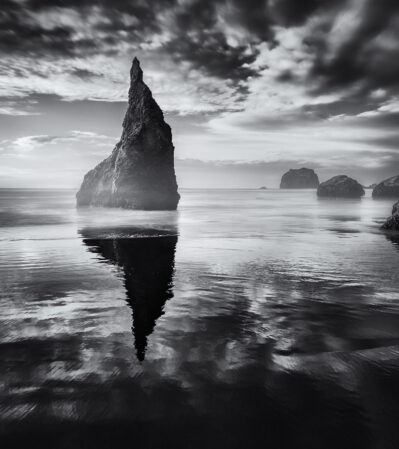 One of the most photographed sea stacks at Bandon Beach, Oregon is Wizard’s Hat. This towering, jagged rock formation rising from the calm water casts a striking reflection on the smooth, mirror-like surface of the ocean, with a symmetrical visual effect. The interplay of shadows and light highlights the textures of the rocks and the gentle movement of the water, contributing to this image ‘s tranquil yet majestic atmosphere.