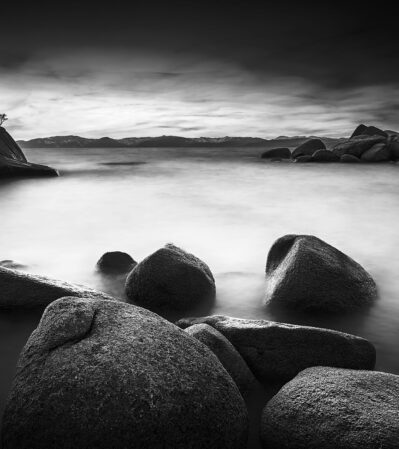 Lake Tahoe is sprinkled with smooth, rounded rocks, immersed partially in misty water. In the middle, there is a larger rock with a couple of small trees growing on top, called Bonsai Rock. The photograph emphasizes textures, contrasts, and a sense of tranquility.