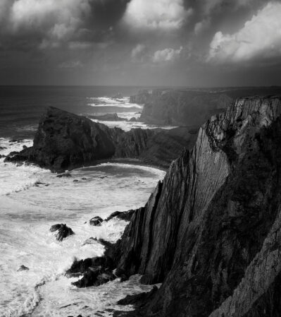 After a spring storm, big waves crush into the dramatic shoreline of Costa Vicentina, Portugal, while the first rays of sun start peeking through the dense clouds.