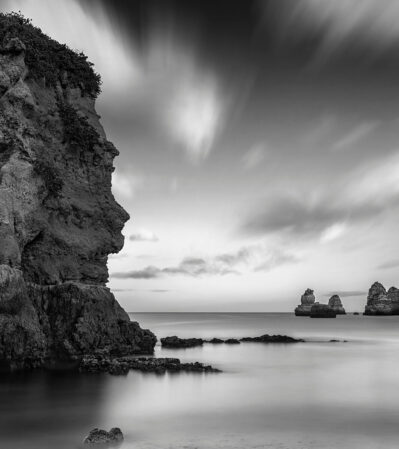 This monochromatic photograph captures a coastal scene at Praia Dona Ana in the Algarve. Dominating the foreground is a large, rugged rock formation emerging majestically from the smooth, reflective surface of the water. The sky is filled with soft, wispy clouds, adding a sense of movement and depth to the composition.