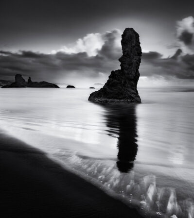 A tall, jagged rock rising from the ocean and its reflection beautifully mirrored in the wet sand below are the subject of this image from Bandon Beach, Oregon. The scene exudes a serene yet powerful atmosphere, with soft, blurred waves washing gently over the beach. The clouds surround and mirror the shape of the sea stack.