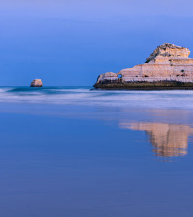 One evening in the Algarve, on Praia da Rocha, I came across a serene coastal scene with two sea stacks emerging from the water reflected beautifully in the calm, glass-like ocean. The overall color palette is composed of soothing blues and gentle purples, creating a tranquil and peaceful mood.