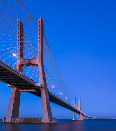 Vasco da Gama Bridge in Lisbon, Portugal connects the two sides separated by the Tagus river. At sunset, the bridge sparkles over the water, bathed in the soft light of the setting sun, and the lights expertly positioned to highlight its brilliant modern architecture.