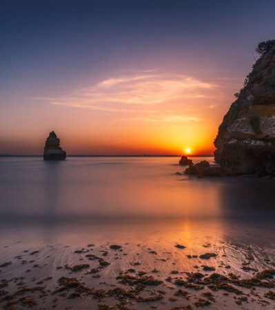 This serene coastal landscape at Ponta da Piedade catches fire when the sun starts to rise above the horizon, casting a golden hue across the scene. The secluded beach in the foreground, with its gentle waves and scattered seaweed, adds texture to the overall tranquil atmosphere of the image.