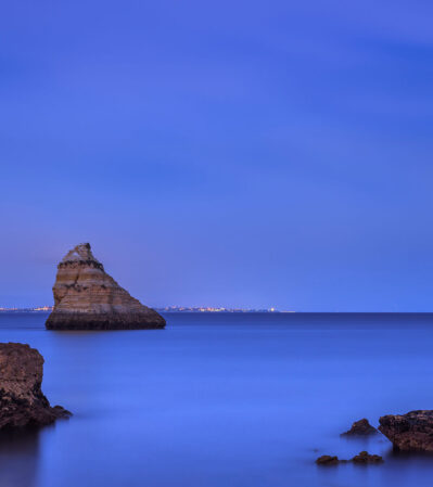 The serene, rich blue hues of the sky beautifully blend with the smooth, calm waters, creating a sense of peace and serenity at Praia Dona Ana in Lagos, Portugal. The foreground is dominated by three rock formations rising from the sea, while distant city lights lining the horizon add a subtle contrast without distracting from the natural beauty of the scene.