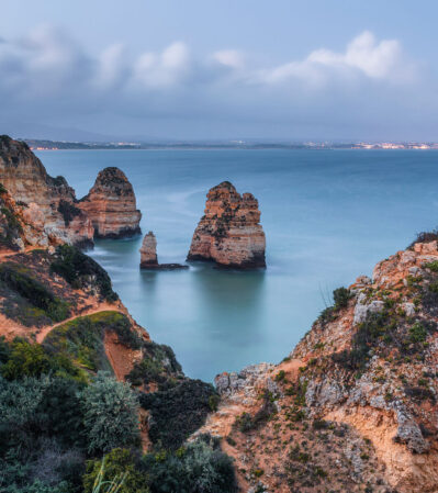 The panoramic view of the coastline at Ponta da Piedade is spectacular. The rugged, weather-worn cliffs and the striking rock formations emerging from the calm, blue waters of the Atlantic ocean contrast with the natural elements, the remains of few storm clouds. The soft light at dusk adds a gentle, almost ethereal quality to the image.
