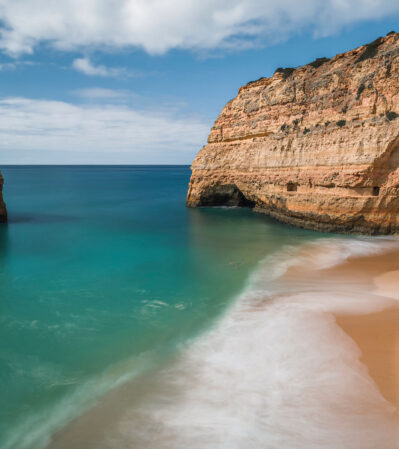 In this serene coastal scene, a stretch of sandy beach bordered by steep, rugged cliffs meets the calm, turquoise sea. Gentle waves, creating a soft, mist-like effect, touch the sand. Off the coastline, a solitary rock rises from the water, adding a striking element to the composition. This serene moment was photographed on a small beach near Carvoeiro, Algarve, Portugal.