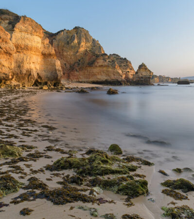 This serene coastal landscape at Ponta da Piedade near Lagos, Portugal features rugged cliffs bathed in warm, golden light, contrasting with the smooth, tranquil water of the sea. The sandy beach in the foreground is peppered with seaweed and rocks, leading the viewer's eye toward the impressive sea stacks.