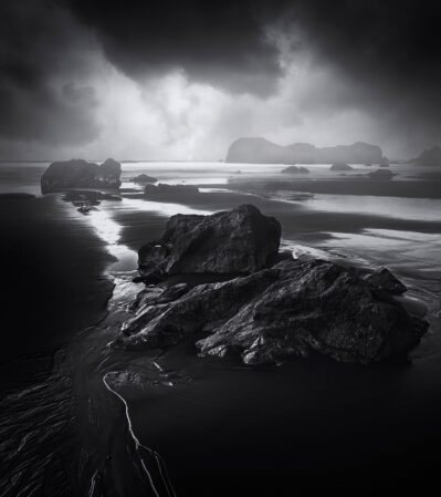 The Pacific coast offers some of the most dramatic landscapes. At Bandon Beach, Oregon, large, rugged rocks are scattered across the shoreline, extending into the water. The wet sand reflects the brooding, overcast sky above. The mood is intense, atmospheric, and slightly ominous, evoking a sense of grandeur and solitude.