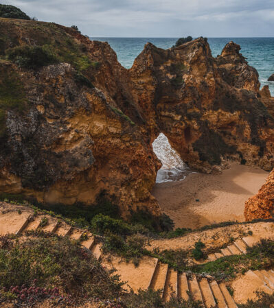 This photograph captures a stunning coastal landscape in the Algarve, Portugal, featuring naturally eroded rock formations with archways overlooking a sandy cove beneath. The rugged cliffs near Portimao, colored in rich earthy tones, contrast beautifully with the deep blue of the ocean. A series of spectacular arches carved into rock extend from the beach into the ocean.